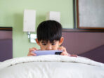 A little Asian boy lies on the bed playing with his phone.